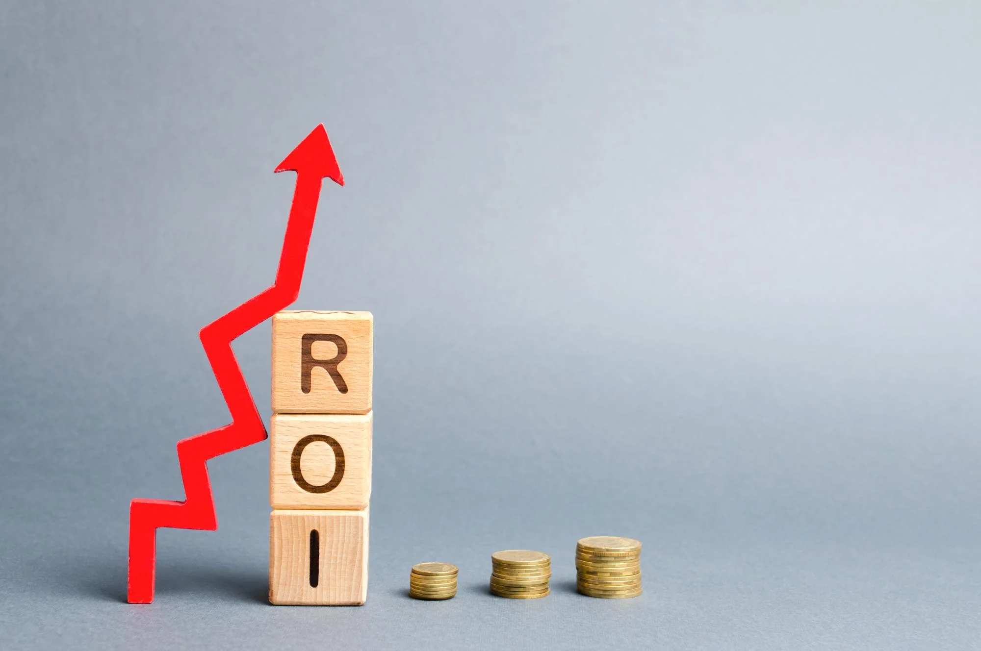 Wooden blocks spelling ROI with a red upward growth arrow and coin stacks representing investment returns